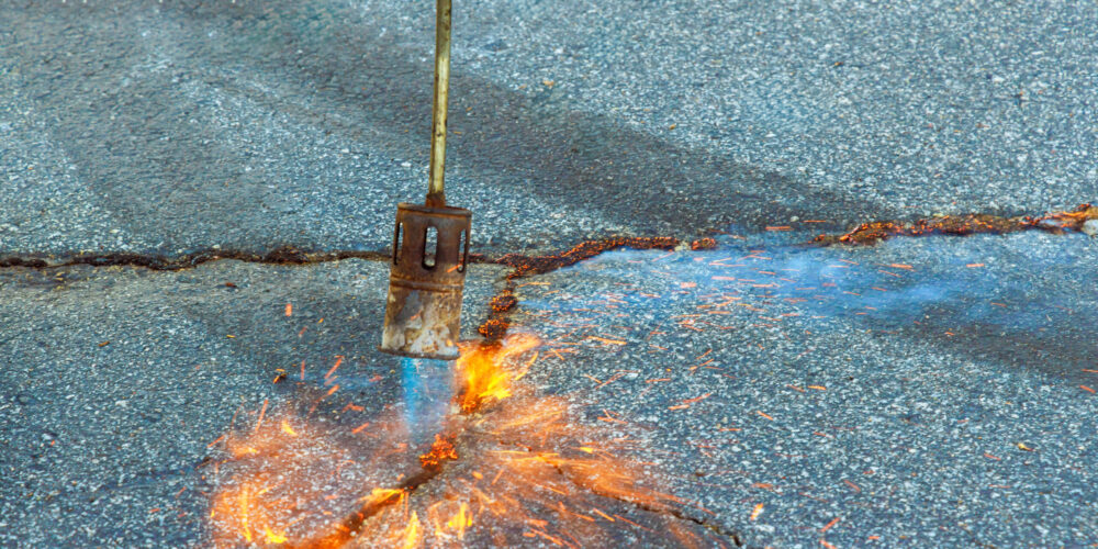 Asphalt patcher performs restoration work in cracks of road surface by burning dry grass before applying coating of bitumen emulsion