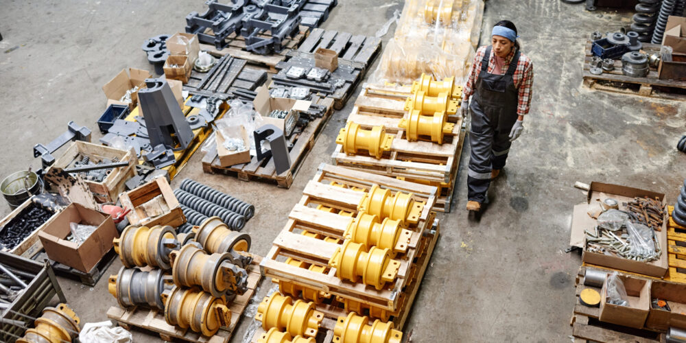 Young female specialist of machine repair service passing by stacks of spare parts while moving along distribution warehouse